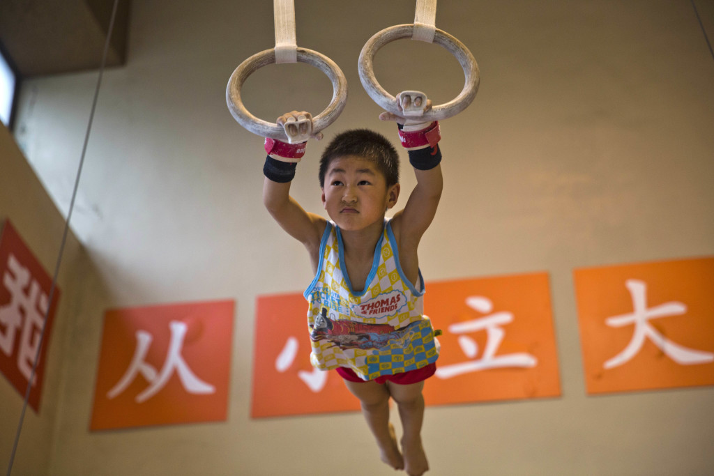 In this photo taken Thursday, June 16, 2016, a young child practices on still rings at the Xuhui Sports School near the slogans "Set ambitious goals from young" in Shanghai, China. The Xuhui Sports School is representative of the state-led sports training system established in the 1950s to churn out hundreds of Olympic gold medalists and world champions but increasingly voices of criticism have grown decrying the state system for its notoriously ruthless, rigid training regimes, exploitation of young athletes and proclivity for dishonest practices such as game rigging. (AP Photo/Ng Han Guan)