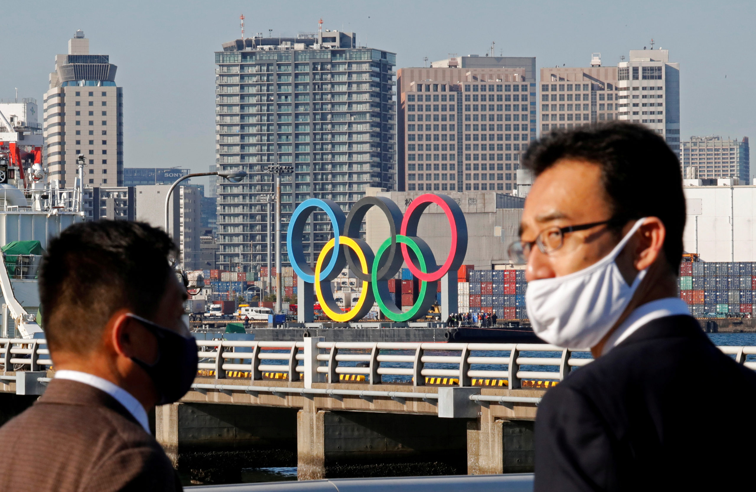 Men wearing face masks watch as giant Olympic rings, which were temporarily taken down in August for maintenance amid the coronavirus disease (COVID-19) outbreak, are transported for reinstallation at the waterfront area at Odaiba Marine Park in Tokyo, Japan December 1, 2020. REUTERS/Kim Kyung-Hoon