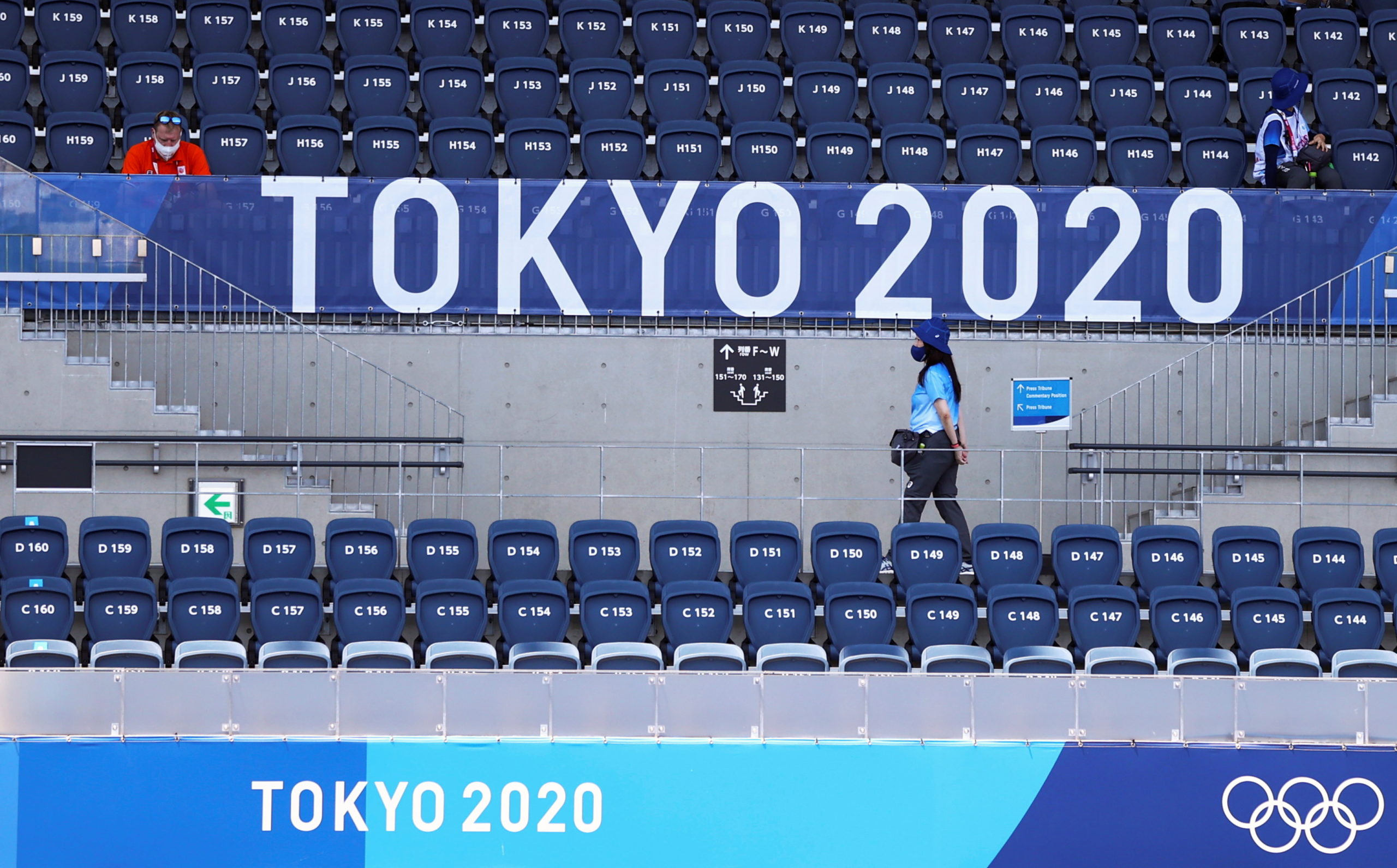 FILE PHOTO: A spectator sits as volunteers walk beneath the logo during the Tokyo 2020 Olympic Games at Oi Hockey Stadium in Tokyo, Japan, July 24, 2021.