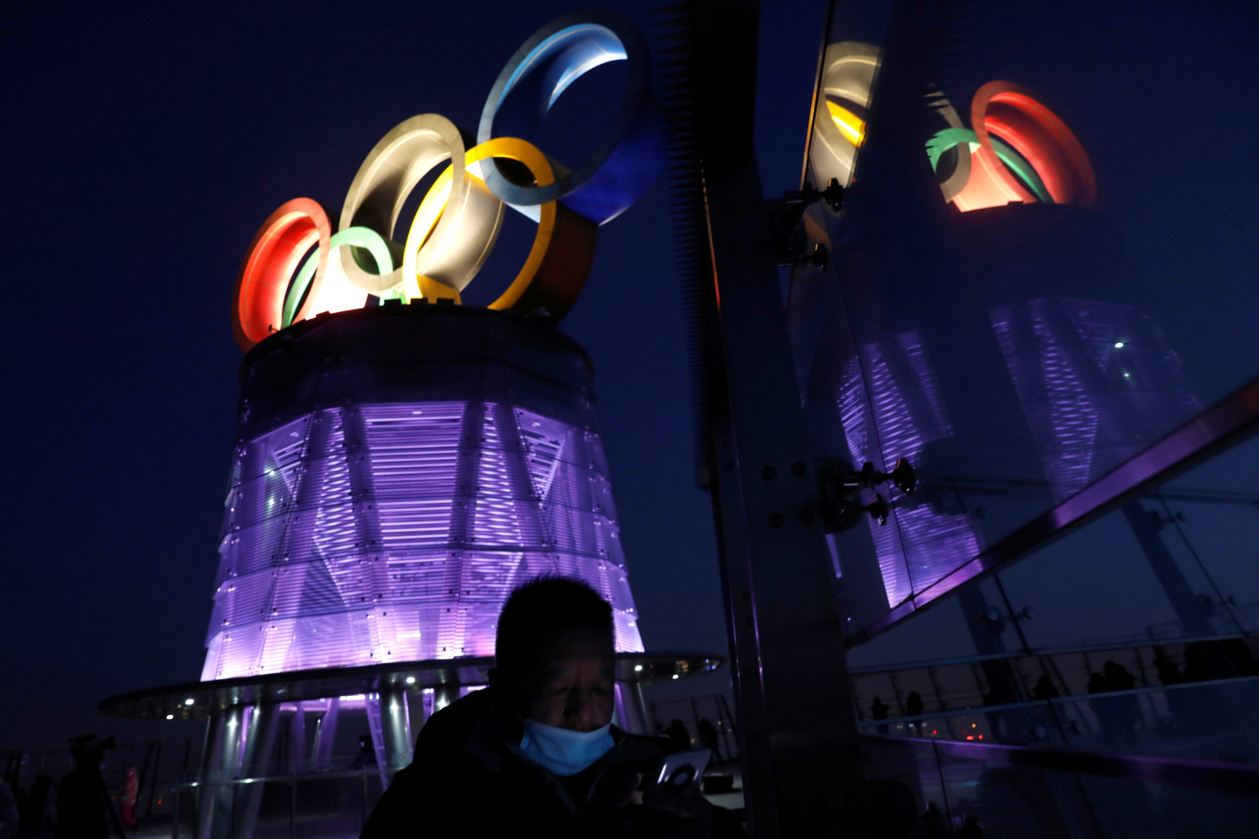 FILE PHOTO: A man wearing a face mask following the coronavirus disease (COVID-19) outbreak is seen near the lit-up Olympic rings at top of the Olympic Tower, a year ahead of the opening of the 2022 Winter Olympic Games, in Beijing, China February 4, 2021. 